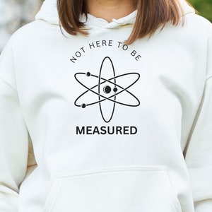 Op de afbeelding: Witte hoodie met een zwarte atoom afbeelding en de tekst "Not here to be measured".
