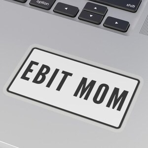 May include: A rectangular white sticker with the text "EBIT MOM" in bold black letters. The sticker is on a silver laptop, near the keyboard. The laptop keyboard is black.