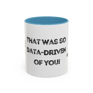 Peut inclure: Une tasse en céramique blanche avec un bord bleu clair. La tasse a un texte noir qui dit "That was so data-driven of you!"