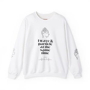 Könnte beinhalten: Weißer Langarm-Pullover mit einem schwarzen Grafikdruck eines Kopfes mit einem Atom darin und dem Text "I wave & particle at the same time" mit einer Herzschlaglinie darunter. Die Ärmel haben eine kleine Grafik eines Atoms und eines Herzens.