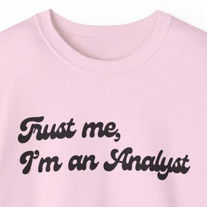 Op de afbeelding: Roze sweatshirt met de tekst "Trust me, I'm an Analyst" in een retro-lettertype.