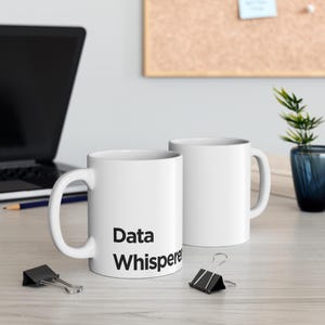 May include: Two white ceramic mugs, one with the text "Data Whisperer" in black lettering. The mugs are on a white surface with a laptop and a pencil in the background.