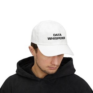 May include: White baseball cap with the text "DATA WHISPERER" in black. The cap is made of a soft material and has a curved brim. The hat is a classic style, suitable for casual wear.