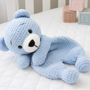 May include: A light blue crochet teddy bear security blanket featuring a textured yarn construction, a white muzzle, black embroidered eyes, and a black nose. The plush toy has a soft, rounded head and a flat, blanket-like body with small limbs.