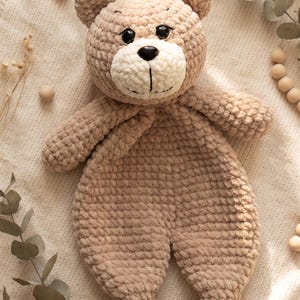 May include: A beige crochet teddy bear lovey with a soft, plush texture. The bear features a round head with two small ears, black plastic eyes, a black plastic nose, and a cream-colored muzzle. The body is flat and blanket-like with two arms.