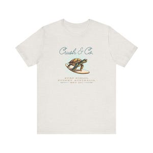 May include: A light grey t-shirt featuring a graphic of a turtle surfing on a surfboard. The text "Crush & Co." is written in cursive above the graphic. Below the graphic is the text "Surf School Sydney Australia Est. 2003."