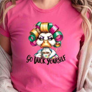 May include: Pink t-shirt featuring a cartoon duck with curlers, holding a coffee cup. The duck has an angry expression and the text "Go Duck Yourself" is below the image. The shirt is a solid pink color.