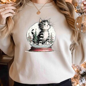 May include: A beige sweatshirt featuring a whimsical illustration of a cat inside a snow globe. The cat is black and white, surrounded by miniature Christmas trees and falling snow. The snow globe sits on a red base, evoking a festive holiday theme.
