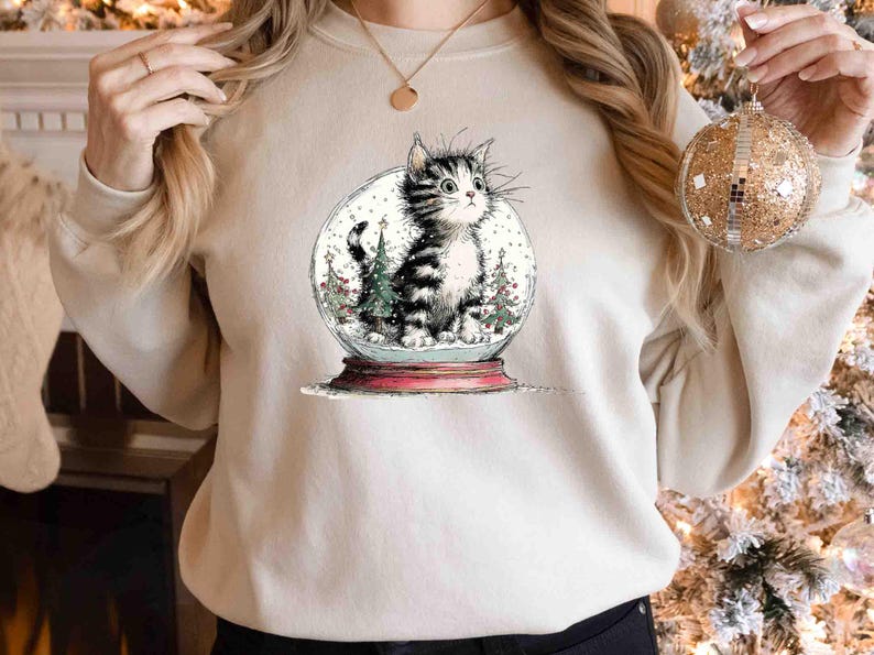 May include: A beige sweatshirt featuring a whimsical illustration of a cat inside a snow globe. The cat is black and white, surrounded by miniature Christmas trees and falling snow. The snow globe sits on a red base, evoking a festive holiday theme.