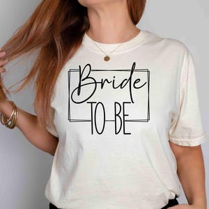K&ouml;nnte beinhalten: Cremefarbenes T-Shirt mit dem Schriftzug "Bride TO BE" in schwarzer Schrift und Blockbuchstaben in einem schwarzen Rechteck. Das Shirt ist im l&auml;ssigen Stil gehalten, passend f&uuml;r eine Hochzeitsfeier.