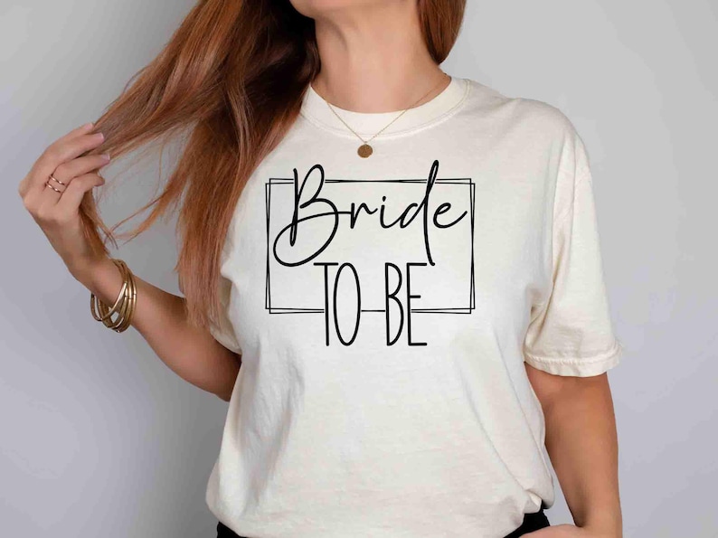 K&ouml;nnte beinhalten: Cremefarbenes T-Shirt mit dem Schriftzug "Bride TO BE" in schwarzer Schrift und Blockbuchstaben in einem schwarzen Rechteck. Das Shirt ist im l&auml;ssigen Stil gehalten, passend f&uuml;r eine Hochzeitsfeier.