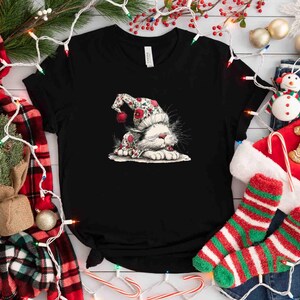 May include: Black t-shirt featuring a whimsical illustration of a white cat wearing a festive floral hat with red accents. The cat is lying down, creating a cozy, holiday-themed design. Christmas-themed apparel.