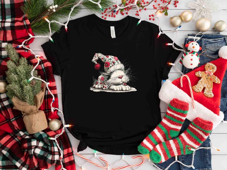May include: Black t-shirt featuring a whimsical illustration of a white cat wearing a festive floral hat with red accents. The cat is lying down, creating a cozy, holiday-themed design. Christmas-themed apparel.
