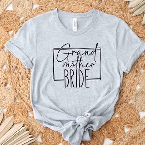 K&ouml;nnte beinhalten: Hellgraues T-Shirt mit dem schwarzen Schriftzug "Grandmother Bride" in Schreib- und Blockschrift in einem schwarzen Rechteck. Das Shirt ist am Saum geknotet und liegt auf einem gewebten, beigefarbenen Teppich.