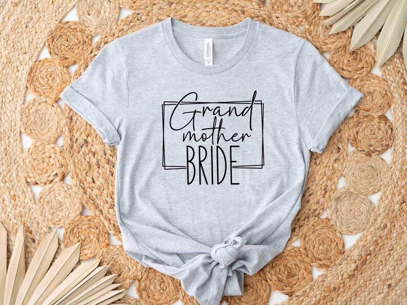 K&ouml;nnte beinhalten: Hellgraues T-Shirt mit dem schwarzen Schriftzug "Grandmother Bride" in Schreib- und Blockschrift in einem schwarzen Rechteck. Das Shirt ist am Saum geknotet und liegt auf einem gewebten, beigefarbenen Teppich.