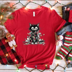 May include: A red t-shirt featuring a black cat wrapped in Christmas lights. The cat has large, wide eyes. The shirt is surrounded by Christmas decorations, including a stocking, socks, and a gift box.