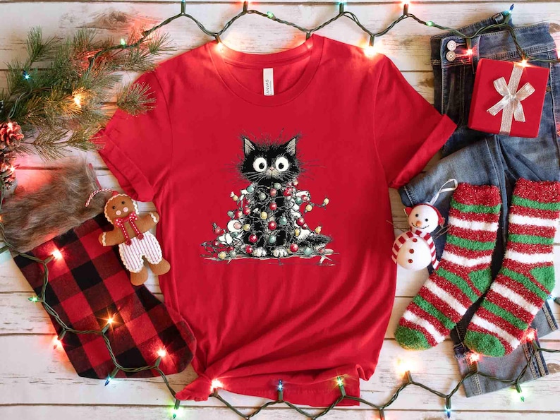 May include: A red t-shirt featuring a black cat wrapped in Christmas lights. The cat has large, wide eyes. The shirt is surrounded by Christmas decorations, including a stocking, socks, and a gift box.