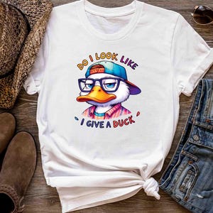 May include: White t-shirt featuring a cartoon duck wearing glasses and a cap, with the text "Do I look like I give a duck". The duck has an orange beak and is wearing a colorful shirt. The shirt is styled with a knot at the bottom.