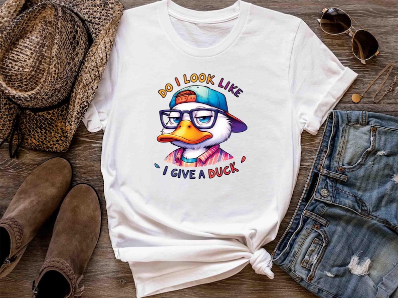 May include: White t-shirt featuring a cartoon duck wearing glasses and a cap, with the text "Do I look like I give a duck". The duck has an orange beak and is wearing a colorful shirt. The shirt is styled with a knot at the bottom.