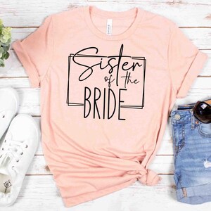 K&ouml;nnte beinhalten: Ein hellrosa T-Shirt mit dem Text "Sister of the Bride" in schwarzer Schrift in einem schwarzen rechteckigen Rahmen. Das Shirt wird mit wei&szlig;en Turnschuhen und Jeansshorts kombiniert, mit Sonnenbrille.