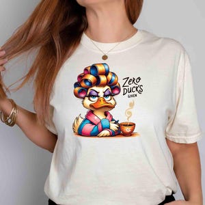 May include: Cream-colored t-shirt featuring a cartoon duck with curlers, a robe, and a cup of coffee. The duck has an annoyed expression. Text on the shirt reads "Zero Ducks Given."
