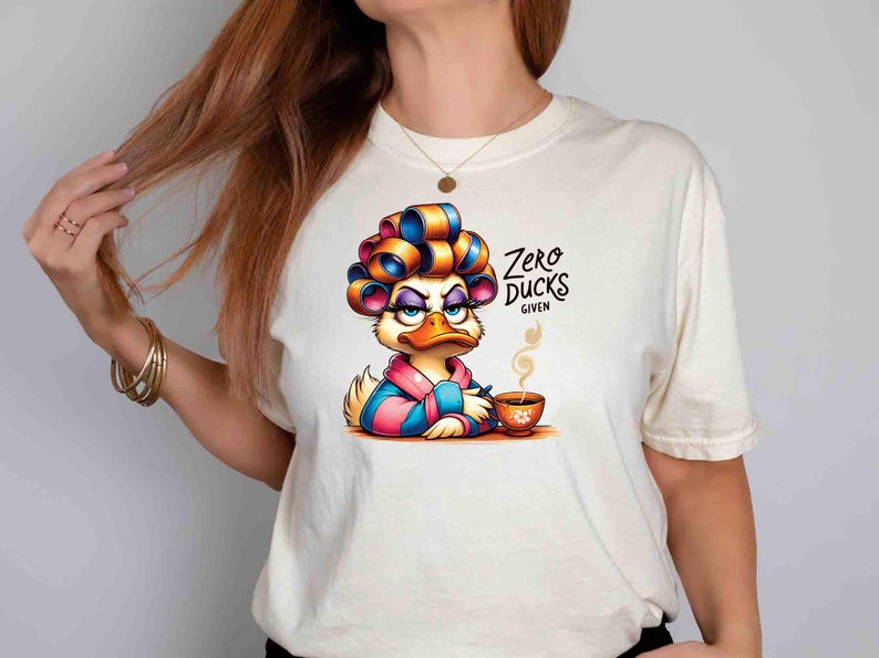 May include: Cream-colored t-shirt featuring a cartoon duck with curlers, a robe, and a cup of coffee. The duck has an annoyed expression. Text on the shirt reads "Zero Ducks Given."