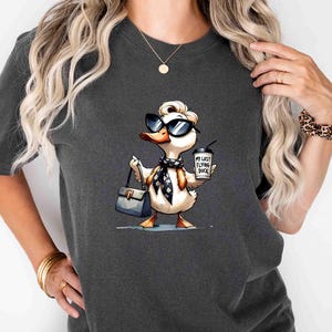 May include: Dark gray t-shirt featuring a cartoon duck wearing sunglasses, a scarf, and carrying a purse and coffee cup. The coffee cup reads "My Last Flying Duck". The duck is a whimsical design.