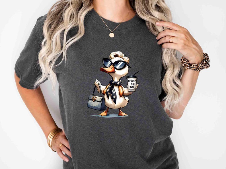 May include: Dark gray t-shirt featuring a cartoon duck wearing sunglasses, a scarf, and carrying a purse and coffee cup. The coffee cup reads "My Last Flying Duck". The duck is a whimsical design.