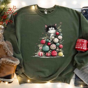 May include: Olive green sweatshirt featuring a whimsical illustration of a black and white cat nestled within a Christmas tree adorned with colorful ornaments. A red mug with a festive pattern and winter boots are also visible.