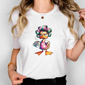May include: White t-shirt featuring a cartoon duck with curlers, wearing a pink heart-patterned apron, and holding a coffee mug. The mug reads "Zero Bucks Given". The duck has an annoyed expression.