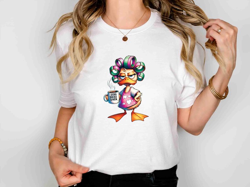 May include: White t-shirt featuring a cartoon duck with curlers, wearing a pink heart-patterned apron, and holding a coffee mug. The mug reads "Zero Bucks Given". The duck has an annoyed expression.