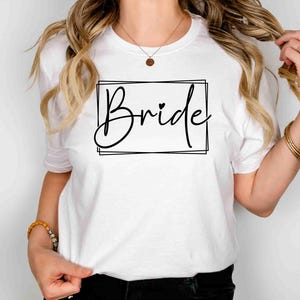 K&ouml;nnte beinhalten: Wei&szlig;es T-Shirt mit dem Wort "Bride" in schwarzer Schreibschrift in einem schwarzen rechteckigen Rahmen. Ein kleines Herz befindet sich &uuml;ber dem "i" in "Bride".
