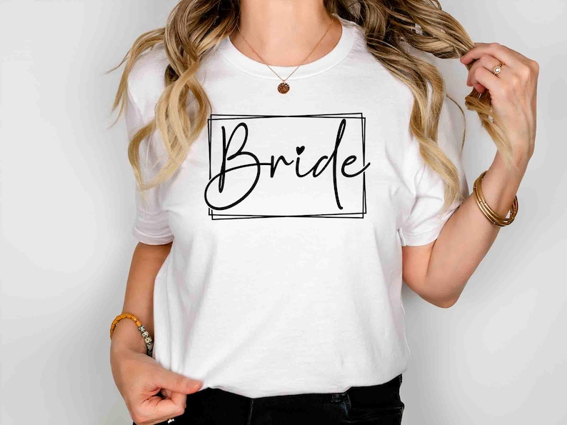 K&ouml;nnte beinhalten: Wei&szlig;es T-Shirt mit dem Wort "Bride" in schwarzer Schreibschrift in einem schwarzen rechteckigen Rahmen. Ein kleines Herz befindet sich &uuml;ber dem "i" in "Bride".