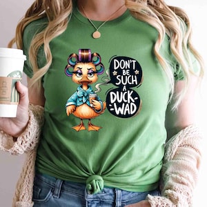 May include: Green t-shirt featuring a cartoon duck with curlers, a blue bow, and a coffee cup. The duck is saying "Don't be such a duck-wad" in a speech bubble. The shirt is a solid green color.
