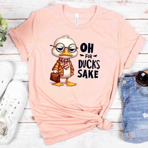 May include: A light pink t-shirt featuring a cartoon duck wearing glasses, a jacket, and a briefcase. The text "OH FOR DUCKS SAKE" is printed on the shirt. The shirt is paired with white sneakers and denim shorts.