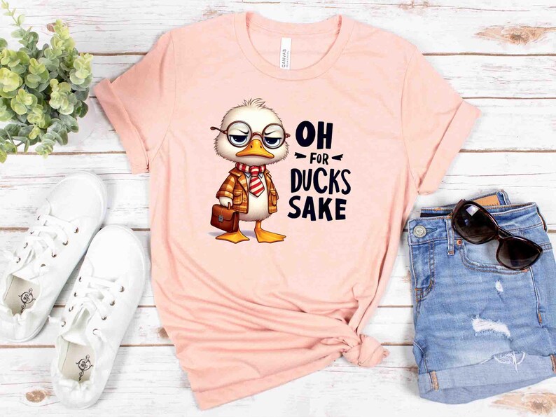 May include: A light pink t-shirt featuring a cartoon duck wearing glasses, a jacket, and a briefcase. The text "OH FOR DUCKS SAKE" is printed on the shirt. The shirt is paired with white sneakers and denim shorts.