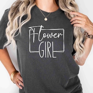K&ouml;nnte beinhalten: Dunkelgraues T-Shirt mit dem wei&szlig;en Schriftzug "Flower Girl" in einem wei&szlig;en rechteckigen Rahmen. Das Shirt hat kurze &Auml;rmel und einen Rundhalsausschnitt. Der Stil ist l&auml;ssig und entspannt.