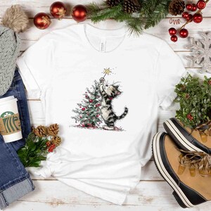 May include: White t-shirt featuring a whimsical illustration of a cat decorating a Christmas tree with a star. The cat is black and gray, and the tree is adorned with red ornaments. The shirt is surrounded by holiday decorations.
