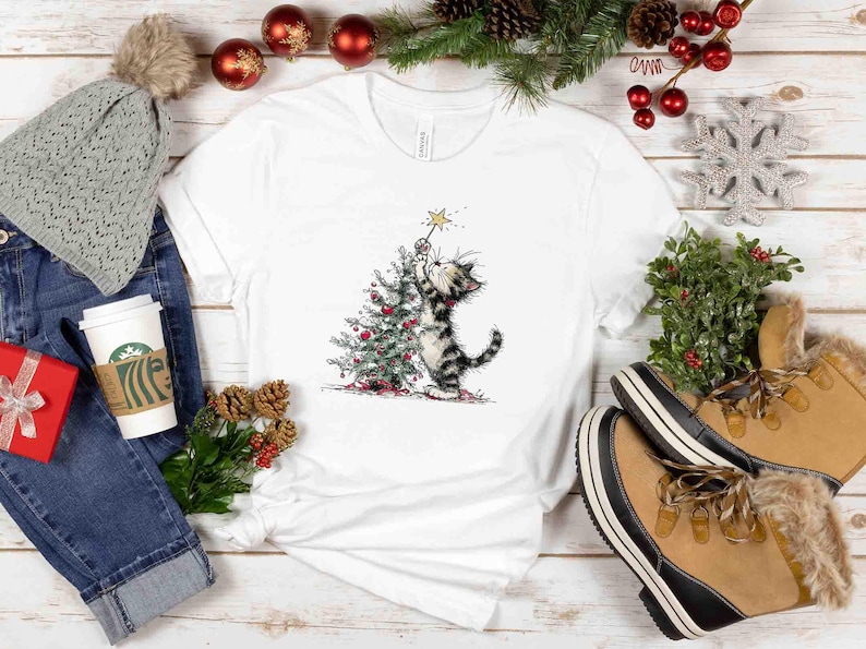 May include: White t-shirt featuring a whimsical illustration of a cat decorating a Christmas tree with a star. The cat is black and gray, and the tree is adorned with red ornaments. The shirt is surrounded by holiday decorations.