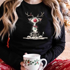 May include: Black long-sleeve shirt featuring a whimsical illustration of a cat dressed as a reindeer with antlers, a red nose, and a green bow tie. The cat is sitting down. A white mug with a holly design is held in the hands.