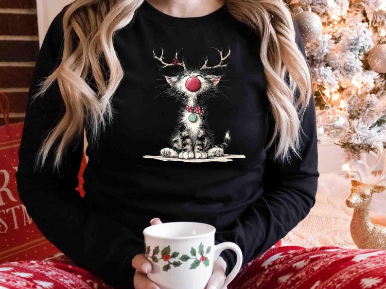 May include: Black long-sleeve shirt featuring a whimsical illustration of a cat dressed as a reindeer with antlers, a red nose, and a green bow tie. The cat is sitting down. A white mug with a holly design is held in the hands.
