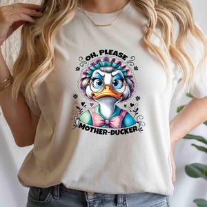 May include: White t-shirt featuring a cartoon duck wearing glasses and a bonnet. The duck has a blue and pink bow tie and the text "OH, PLEASE MOTHER-DUCKER" is printed below the duck image. The shirt is a casual style.