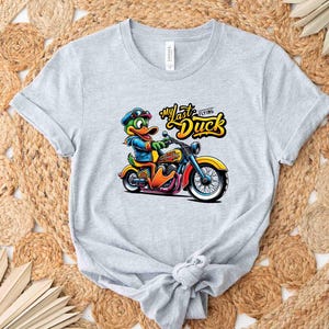 May include: A light gray t-shirt featuring a cartoon duck riding a motorcycle. The duck is wearing a blue jacket, goggles, and a cap. The text "My Last Flying Duck" is above the duck. The motorcycle is yellow, orange, and blue.