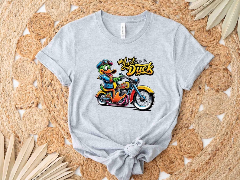 May include: A light gray t-shirt featuring a cartoon duck riding a motorcycle. The duck is wearing a blue jacket, goggles, and a cap. The text "My Last Flying Duck" is above the duck. The motorcycle is yellow, orange, and blue.