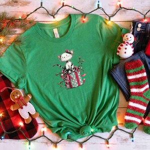 May include: A green t-shirt featuring a cat sitting on a gift box with a bow. The image includes a snowman ornament, red gift box, red and green striped socks, and a gingerbread man ornament. Christmas theme.