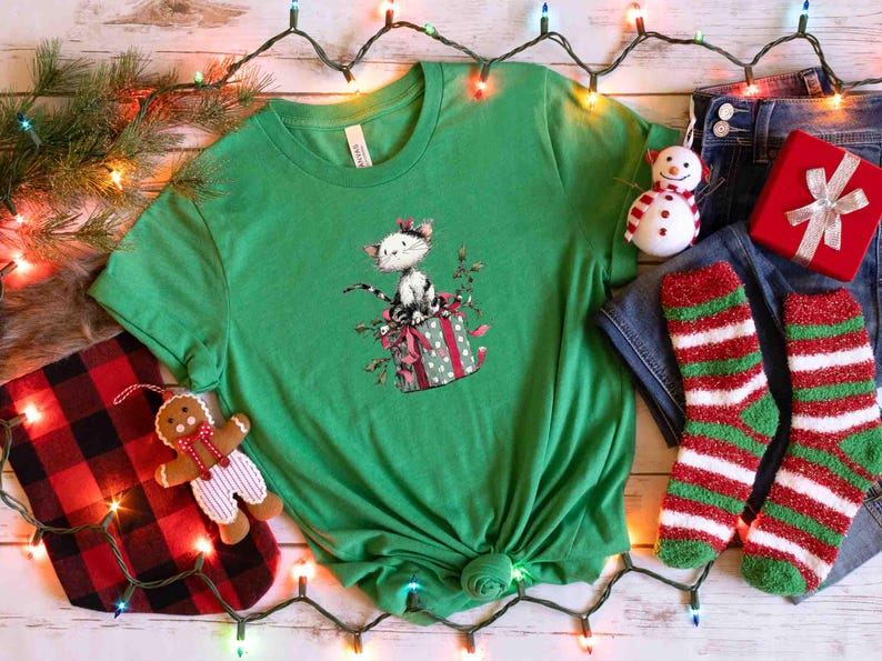 May include: A green t-shirt featuring a cat sitting on a gift box with a bow. The image includes a snowman ornament, red gift box, red and green striped socks, and a gingerbread man ornament. Christmas theme.