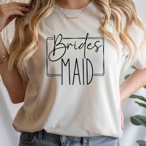 K&ouml;nnte beinhalten: Cremefarbenes T-Shirt mit dem Schriftzug "Bridesmaid" in schwarzer, eleganter Schrift, umrahmt von einem einfachen schwarzen Rechteck. Das Shirt hat einen Rundhalsausschnitt und scheint aus weichem Material zu sein.