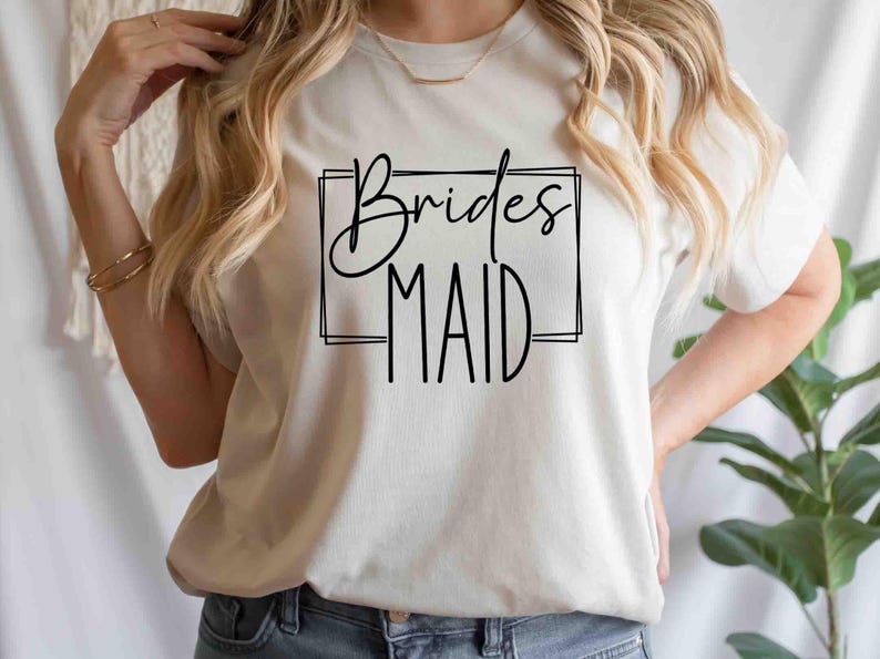 K&ouml;nnte beinhalten: Cremefarbenes T-Shirt mit dem Schriftzug "Bridesmaid" in schwarzer, eleganter Schrift, umrahmt von einem einfachen schwarzen Rechteck. Das Shirt hat einen Rundhalsausschnitt und scheint aus weichem Material zu sein.