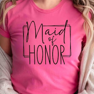 K&ouml;nnte beinhalten: Ein leuchtend pinkfarbenes T-Shirt mit dem schwarzen Schriftzug "Maid of Honor" in einem schwarzen rechteckigen Rahmen. Das T-Shirt hat kurze &Auml;rmel und wird von einer Person mit blonden Haaren getragen.