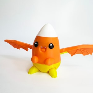 May include: A small orange, yellow, and white candy corn shaped toy with wings. The toy has a cute cartoon face with black eyes and a small mouth.